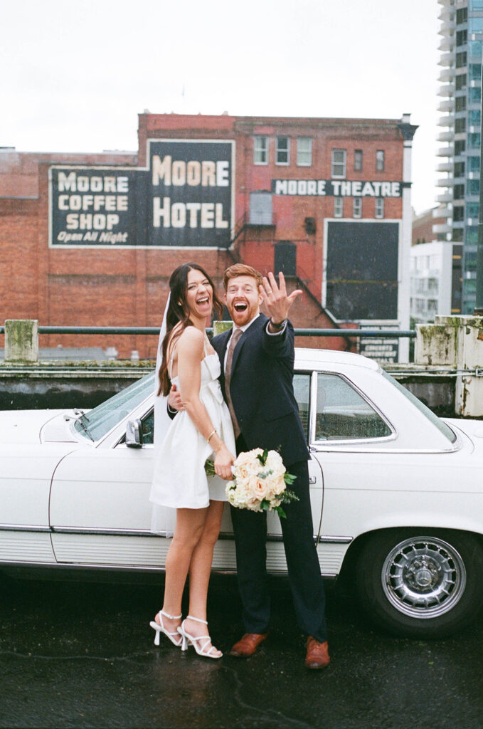 Bride and groom laughing and showing off their rings while standing in front of a vintage white car with Moore Theatre in the background.