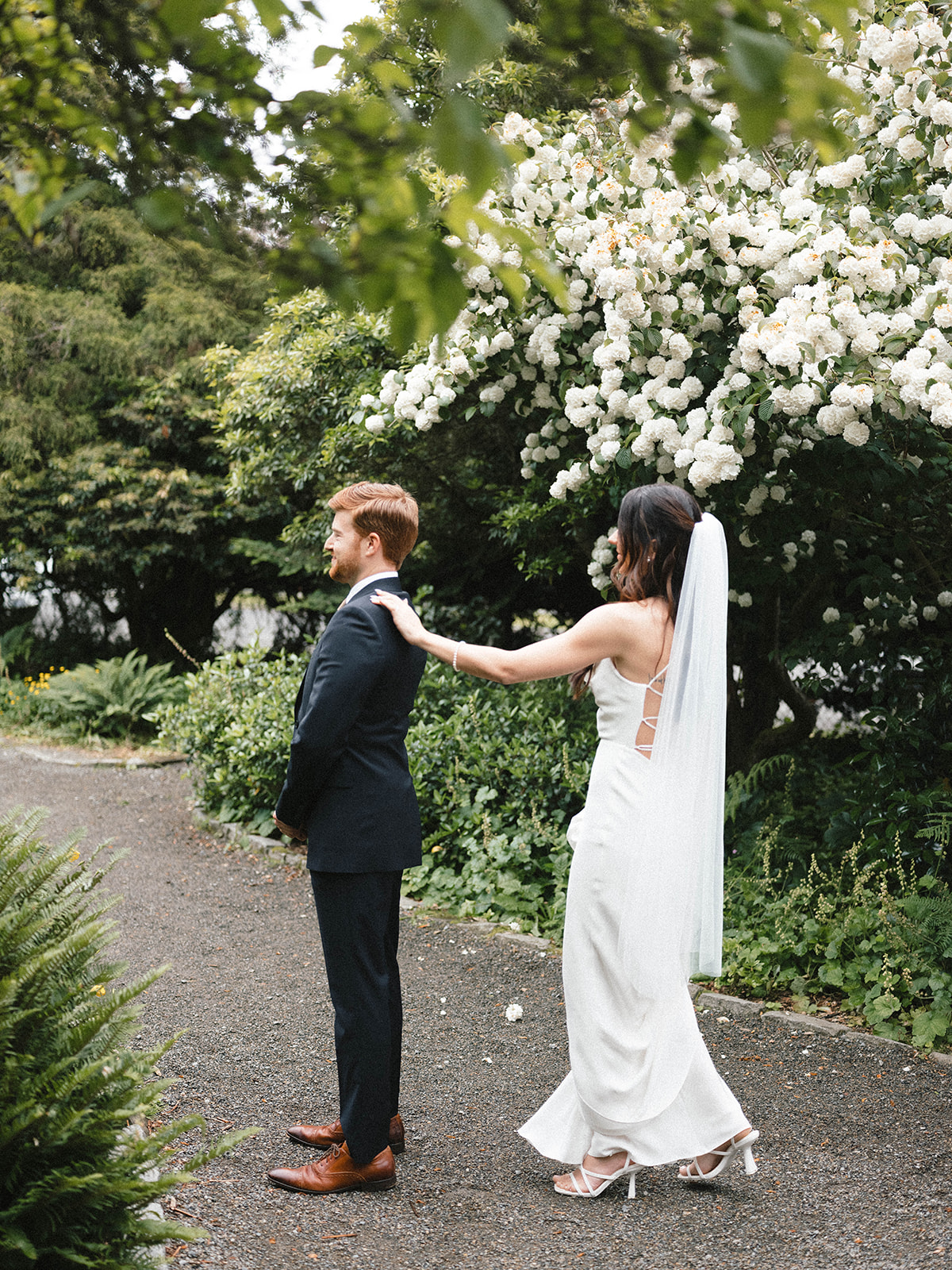 Bride approaches the groom from behind during their first look at Parsons Gardens, surrounded by lush greenery and white flowering shrubs.