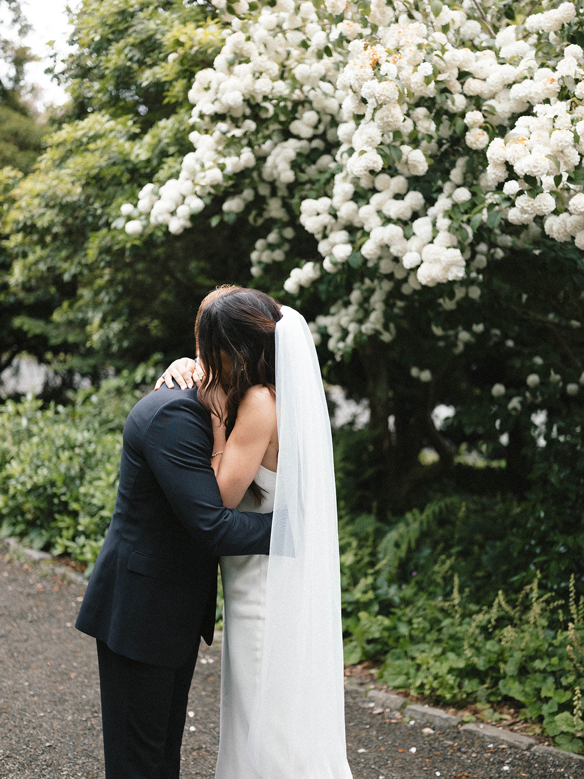 Couple embracing during their first look in a serene garden, surrounded by vibrant greenery and white blossoms.