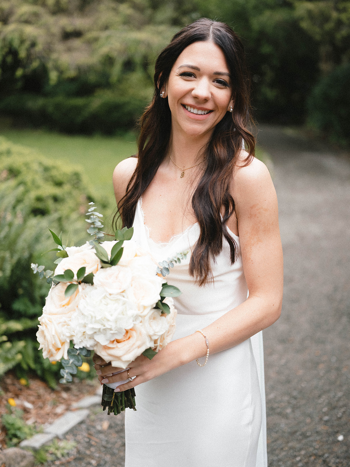 Bride smiling brightly while holding a bouquet of soft white and blush roses in a garden setting.