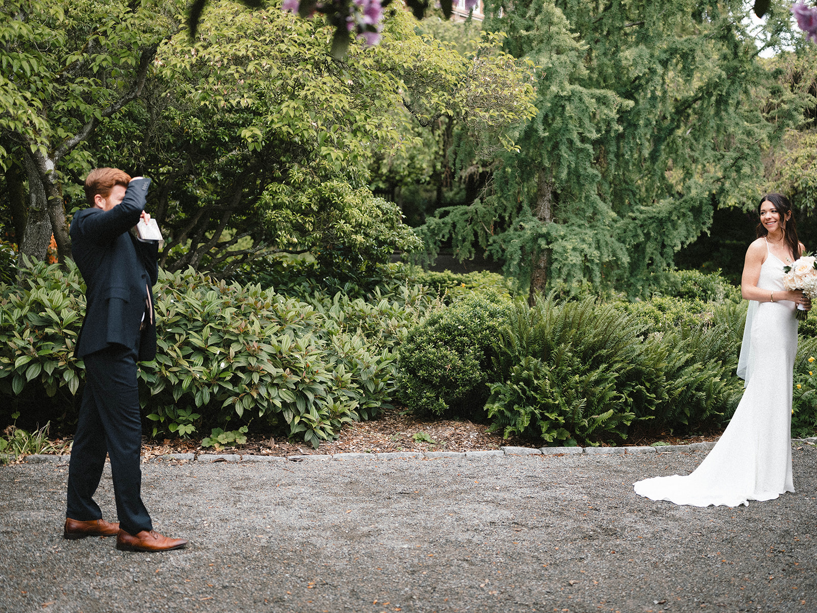 Groom holds camera as the bride smiles during their emotional first look in Parsons Gardens, surrounded by lush greenery.