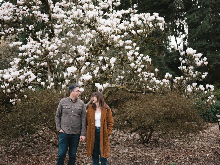 Charlotte and Grant holding hands and smiling in front of a blooming magnolia tree.