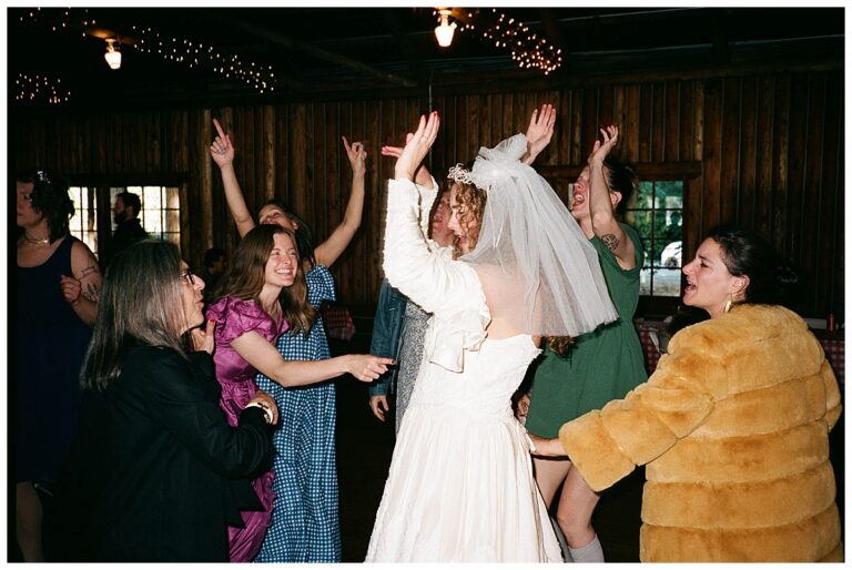 The bride dances joyfully with friends and family, celebrating with laughter and raised hands at the reception.