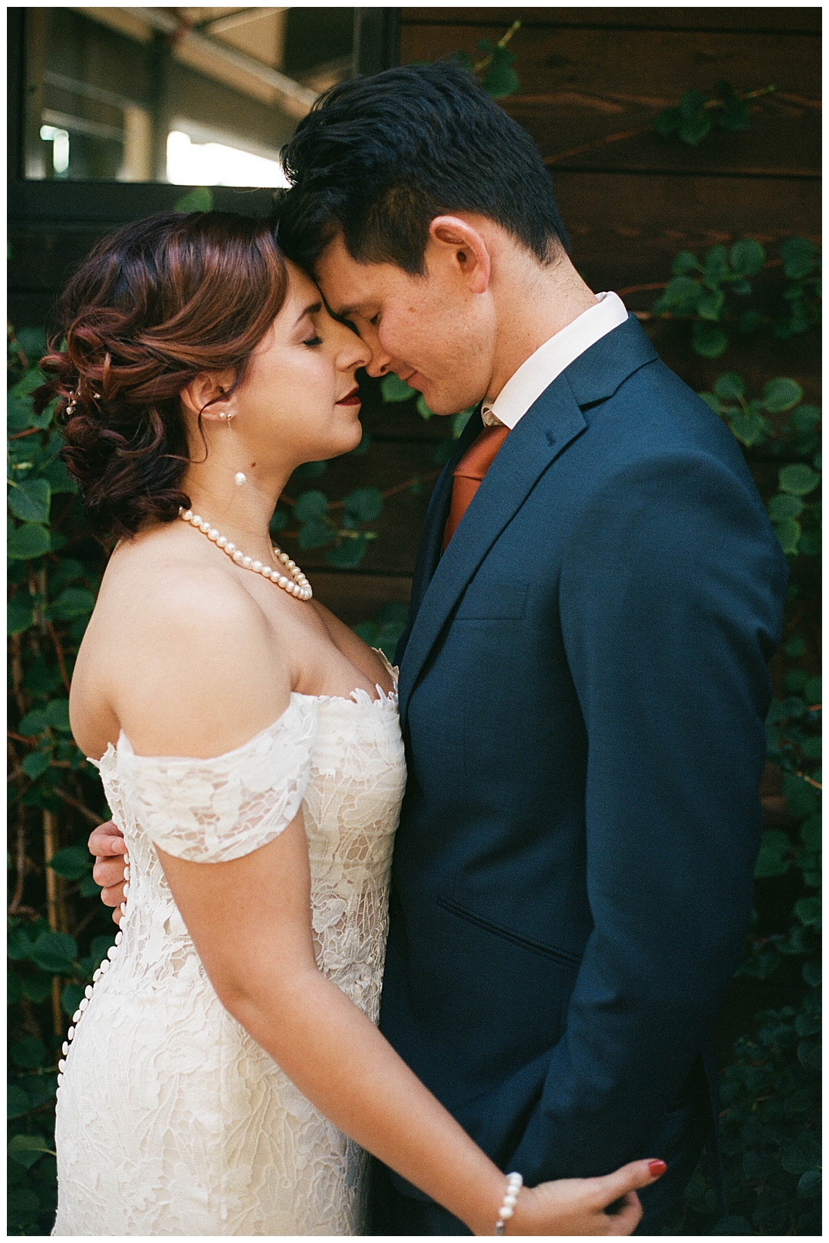 Bride and groom in an intimate embrace, standing against a wooden wall covered in green ivy.