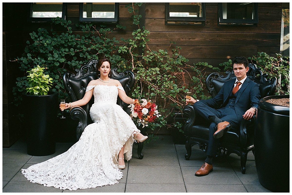 Bride and groom seated in black statement armchairs with a dramatic backdrop of ivy-covered walls.